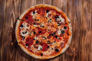 Tomato-based pizza with mozzarella cheese, hunting sausages, salami, olives, mushroom sauce. On a wooden background.