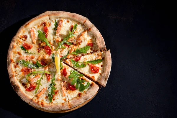 Pizza with salmon, mozzarella, cherry tomatoes, arugula, lemon and parmesan. Italian cuisine. On a black background. Free space for text. View from above