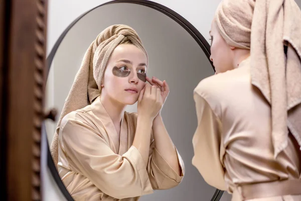 Eye patches for swelling, wrinkles, dark circles. Eye patches. Satisfied woman in cream gown applies brown patches to eyes in bathroom looking in mirror.