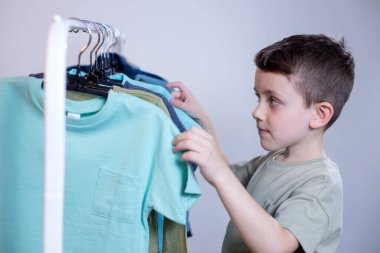 Anaokulu çocuğu askıların yanında duruyor, elbiselerini diziyor ve bugün için kıyafet seçiyor..