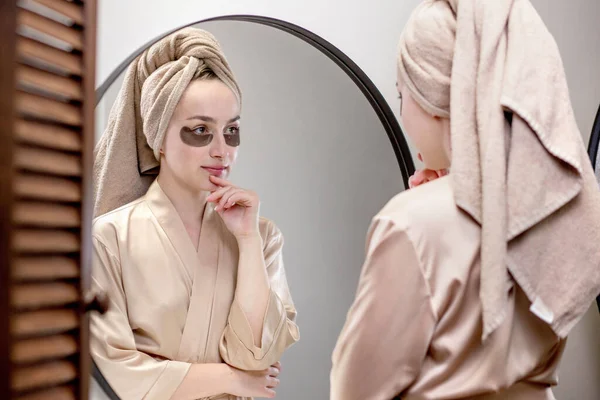 Eye patches for swelling, wrinkles, dark circles. Eye patches. Satisfied woman in cream gown applies brown patches to eyes in bathroom looking in mirror.