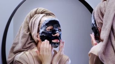 Genç bir kadın banyoda maske takıyor ve gülümsüyor. Güzel bir kadın aynanın önünde güzellik tedavisi yapıyor.