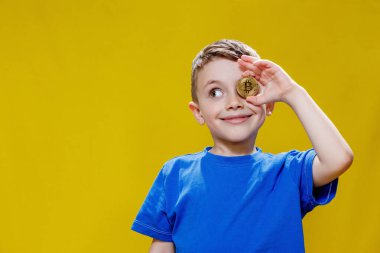 Mavi tişörtlü küçük mutlu anaokulu çocuğu sarı arka planda gözlerinin önünde Bitcoin tutuyor..
