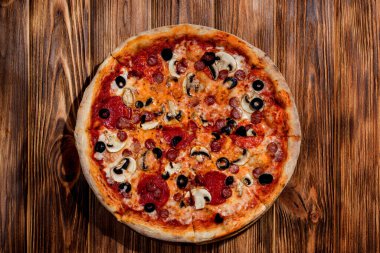 Tomato-based pizza with mozzarella cheese, hunting sausages, salami, olives, mushroom sauce. On a wooden background.