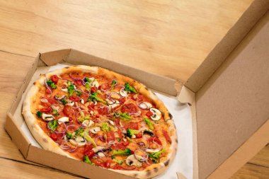 Vegan pizza in a cardboard box on a wooden background. Pizza with mushrooms, onions, broccoli, cherry tomatoes on a tomato base.