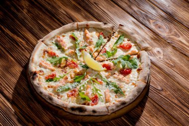 Pizza with salmon, mozzarella, cherry tomatoes, arugula, lemon and parmesan. Italian cuisine on a woden background.