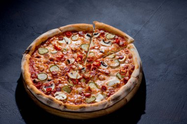 Appetizing pizza with hunting sausages, mozzarella cheese, pickled cucumbers, chili peppers on a tomato base on black background.