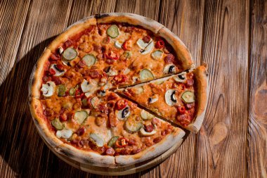 Appetizing pizza with hunting sausages, mozzarella cheese, pickled cucumbers, chili peppers on a tomato base on wood background.