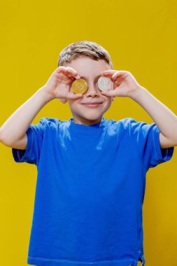 Mavi tişörtlü küçük mutlu anaokulu çocuğu sarı arka planda gözlerinin önünde ethereum ve bitcoin paralar tutuyor..