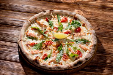 Pizza with salmon, mozzarella, cherry tomatoes, arugula, lemon and parmesan. Italian cuisine on a woden background.
