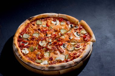 Appetizing pizza with hunting sausages, mozzarella cheese, pickled cucumbers, chili peppers on a tomato base on black background.