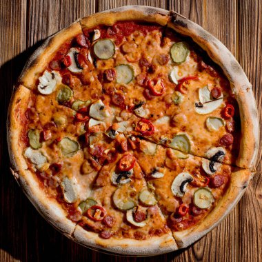 Appetizing pizza with hunting sausages, mozzarella cheese, pickled cucumbers, chili peppers on a tomato base on wood background.