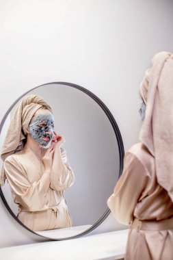 Genç bir kadın banyoda maske takıyor ve gülümsüyor. Güzel bir kadın aynanın önünde güzellik tedavisi yapıyor.