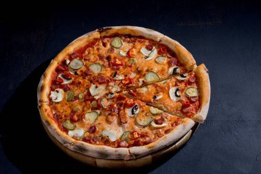 Appetizing pizza with hunting sausages, mozzarella cheese, pickled cucumbers, chili peppers on a tomato base on black background.