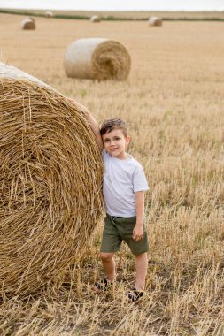 Beyaz tişörtlü ve haki şortlu şirin bir çocuk saman balyalarının yanında duruyor. Sahadaki fotoğraf oturumu.