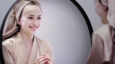 Genç ve mutlu bir kadın, şık bir banyoda aynaya bakarak deriyi temizlemek için maskesini çıkardıktan sonra. Cilt bakımı