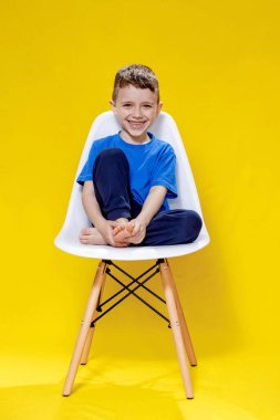 Mavi tişörtlü ve pantolonlu neşeli küçük çocuk sarı arka plan stüdyosunda beyaz bir sandalyede oturuyor..