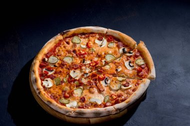 Appetizing pizza with hunting sausages, mozzarella cheese, pickled cucumbers, chili peppers on a tomato base on black background.