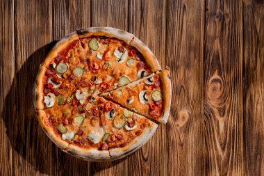Appetizing pizza with hunting sausages, mozzarella cheese, pickled cucumbers, chili peppers on a tomato base on wood background.