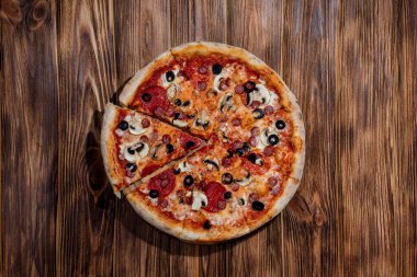 Tomato-based pizza with mozzarella cheese, hunting sausages, salami, olives, mushroom sauce. On a wooden background.