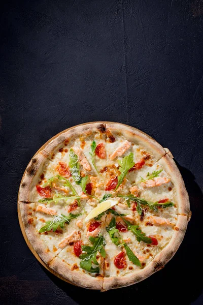 Pizza with salmon, mozzarella, cherry tomatoes, arugula, lemon and parmesan. Italian cuisine. On a black background. Free space for text. View from above