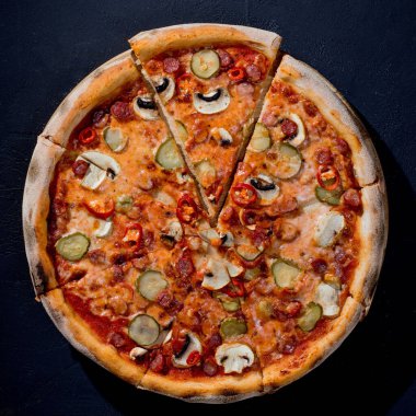 Appetizing pizza with hunting sausages, mozzarella cheese, pickled cucumbers, chili peppers on a tomato base on black background.