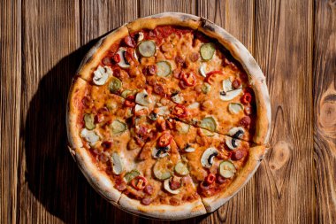 Appetizing pizza with hunting sausages, mozzarella cheese, pickled cucumbers, chili peppers on a tomato base on wood background.