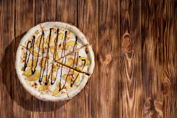 Sweet delicious pizza with apple, cinnamon, chocolate on wood background. Pizza dessert. Top view