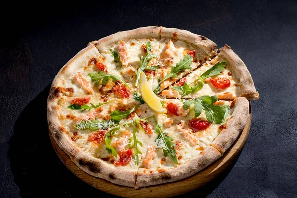Pizza with salmon, mozzarella, cherry tomatoes, arugula, lemon and parmesan. Italian cuisine. On a black background