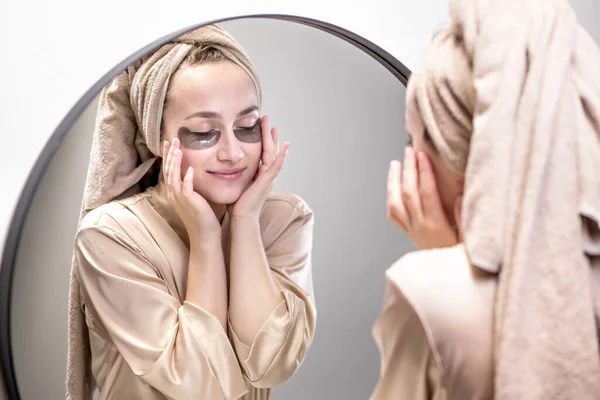 Eye patches for swelling, wrinkles, dark circles. Eye patches. Satisfied woman in cream gown applies brown patches to eyes in bathroom looking in mirror.