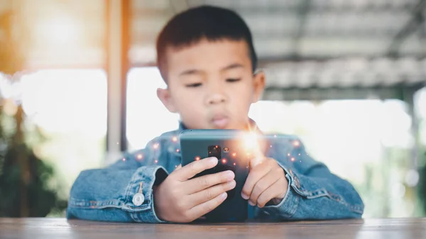 Asian boy looking at the mobile phone screen attentively. overstimulated children concept. Too much screen time. 5 years boy watching videos while tv is working.Smartphone, Internet addiction concept.
