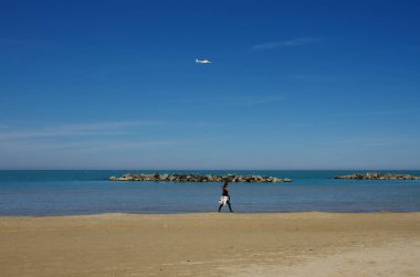 Bir kız mavi gökyüzünde uçan bir helikopter gibi denizin kenarında yürüyor.