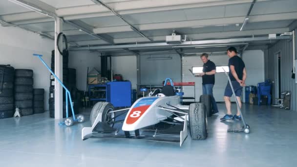 Garage med en racerbil observeras av två mekaniker — Stockvideo