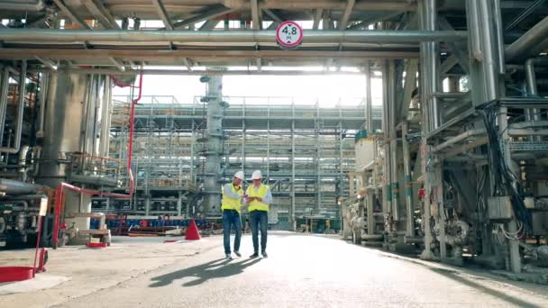 Два инспектора нефтеперерабатывающего завода проходят по трубопроводам во время разговора — стоковое видео
