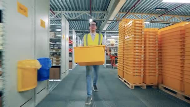 Egy fényvisszaverő mellényes férfi cipel egy dobozt a raktáron keresztül. — Stock videók