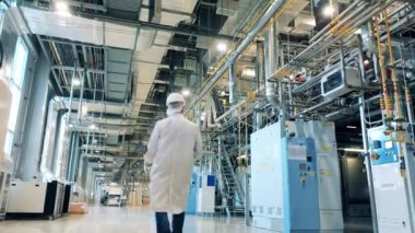 Laboratuvar önlüğü giymiş bir erkek, fabrika makinelerini ve boruları inceliyor.