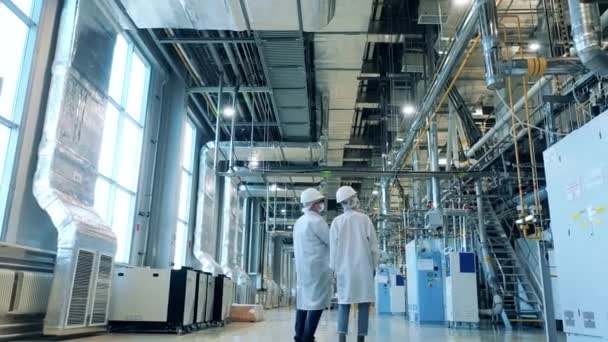 研究室のコートの2人の専門家が工場を歩いている。工場施設、近代的な工業プラントで働く技術者. — ストック動画