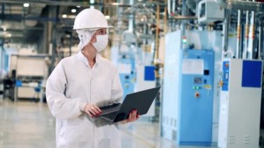 Yüz maskesi takmış bir fabrika mühendisi laptopa veri giriyor.