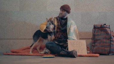 Evsiz bir adam yerde otururken köpeğini okşuyor.