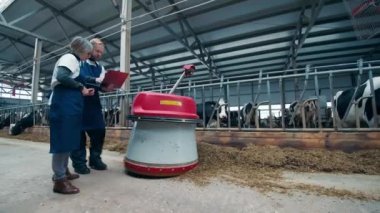 İki uzman, sığır kulübesinde bir robotu gözlemliyor.