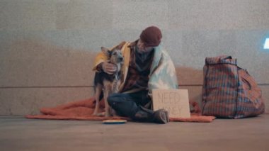 A beggar is stroking his dog while sitting on the ground