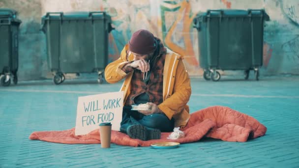 A beggar is eating on the ground near waste bins — Stock video