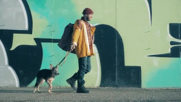 A beggar with his dog are passing by a graffiti wall — Wideo stockowe