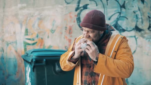 A poor man is eating a hamburger next to the waste bins — стоковое видео