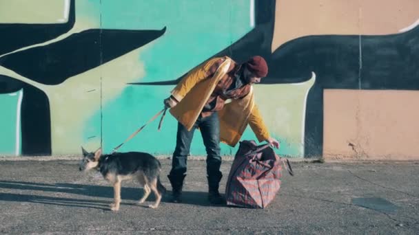 Ένας ζητιάνος στέκεται δίπλα σε έναν τοίχο γκράφιτι με το σκύλο του. — Αρχείο Βίντεο