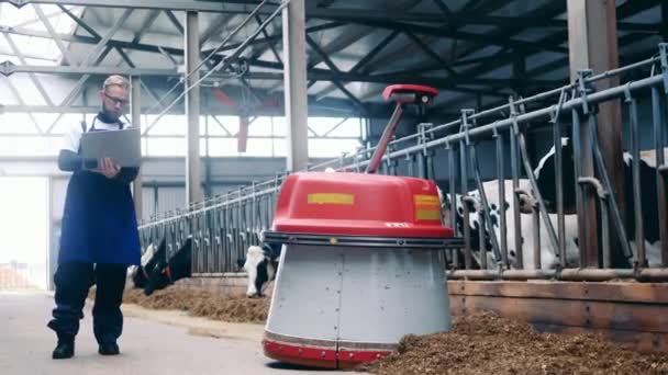 Farm expert with a laptop is following an automated feed pusher — Vídeo de Stock