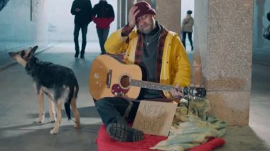 Evsiz bir adam yerde gitar ve köpeğiyle oturuyor.