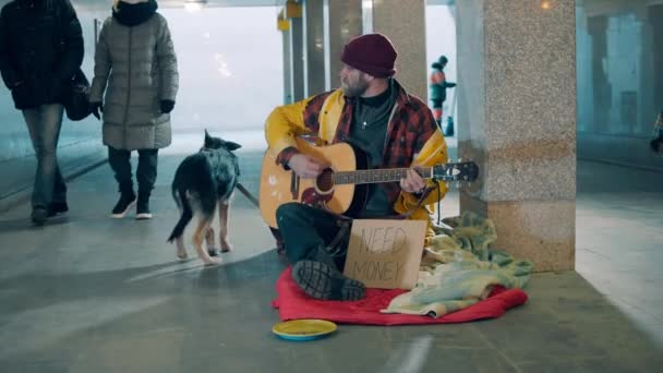 Подающий милостыню играет на гитаре в подземке со своей собакой — стоковое видео