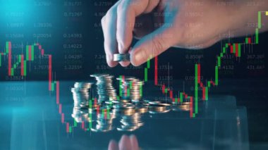 A hand adds coins to a pile with stock charts on the screen
