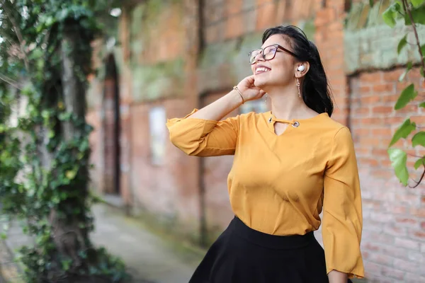 Young Latin American woman wearing eyeglasses and earbuds looking up smiling with leaves copy space. High quality photo